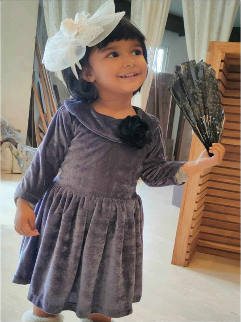 Girl wearing grey 3/4th sleeves velvet party frock with bow attachment and white hair accessory and holding a fan